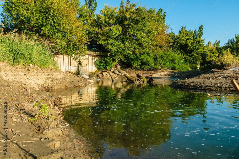Drainage of sewage into the river. A small river with foam on the surface against a background of green trees and blue sky. The concept of ecology. Drainage of sewage into the river. A small river with foam on the surface against a background of green trees and blue sky. The concept of ecology.