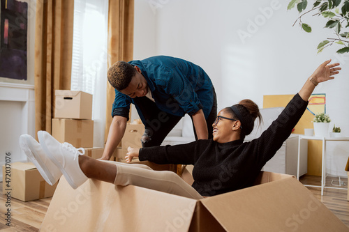 Happy couple first time home buyers having fun while unpacking boxes of laughter on moving day excited wife driving around sitting in cardboard box while husband pushes her around in new apartment photo