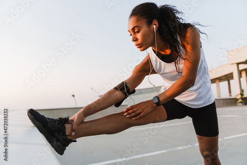 Black sportswoman listening music while doing exercise on parking