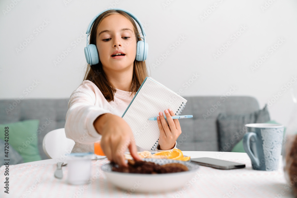 Naklejka premium White girl in headphones writing down notes while having breakfast