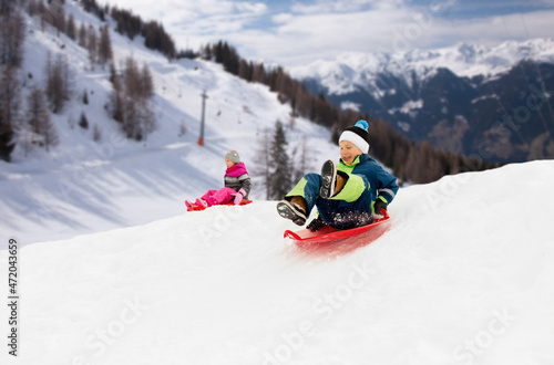 childhood, sledging and season concept - happy little kids sliding on sleds down snow hill in winter over alps mountains on background