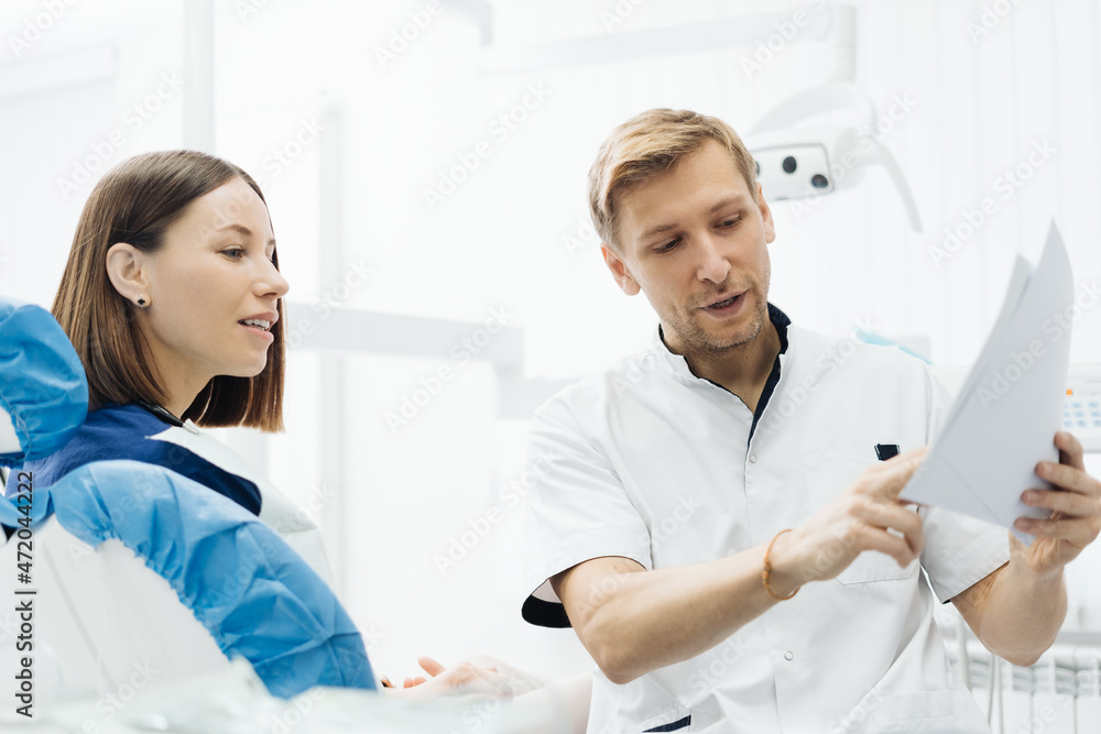 Obraz premium Male Professional Dentist With Gloves And Mask Holding papers photo And Show What The Treatment Will Look Like Of The Patient's Teeth. Discussion Of The Treatment Plan And Healthy Smile