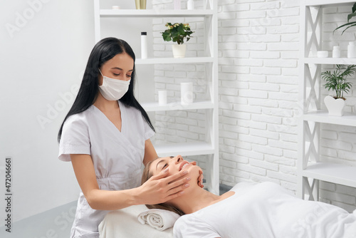 Konstfotografi Side view of young brunette beautician in protective mask doing cleaning and massage face for young beautiful patient