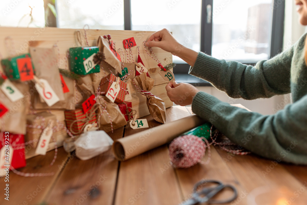 winter holidays, christmas and hobby concept - hands hanging gift with tag to handmade advent calendar at home