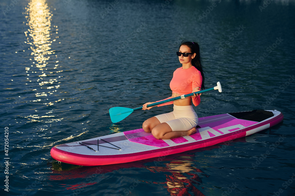 Young caucasian woman in sportswear using long paddle for swimming on sup board on city lake. Reflection from summer sunset on water surface. Outdoors workout. 