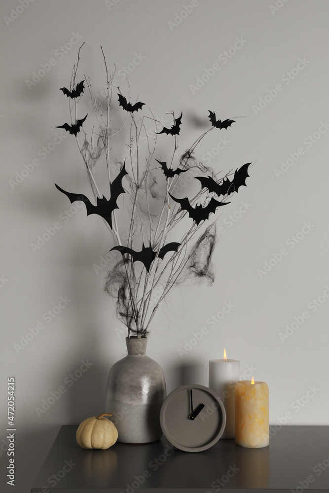 Alarm clock, burning candles and vase with paper bats  on table near white wall. Halloween decor - obrazy, fototapety, plakaty 