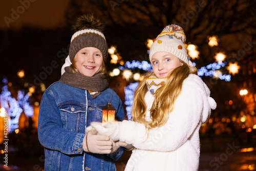 Funny couple of children  girl and boy  hold lantern in their hands against background of christmas lights outside  in warm winter clothes and knitted hats. Christmas and childhood concept  outdoor