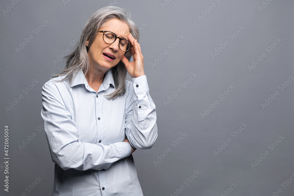 Tired senior mature gray-haired businesswoman holding forehead standing isolated on gray with eyes closed, elder female entrepreneur ceo manager feels headache, suffers from migrain, burnout Tired senior mature gray-haired businesswoman holding forehead standing isolated on gray with eyes closed, elder female entrepreneur ceo manager feels headache, suffers from migrain, burnout