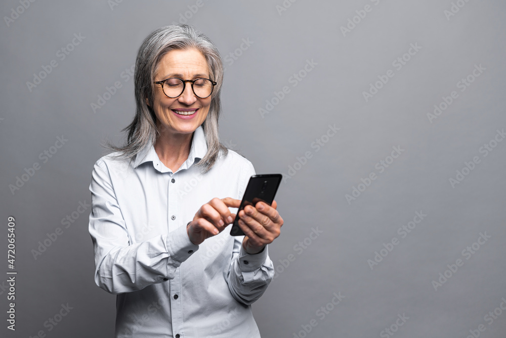 Cheerful positive happy senior gray-haired business woman using smartphone standing isolated on gray, mature lady in formal wear touching touchscreen, scrolling news feed, messaging, texting Cheerful positive happy senior gray-haired business woman using smartphone standing isolated on gray, mature lady in formal wear touching touchscreen, scrolling news feed, messaging, texting