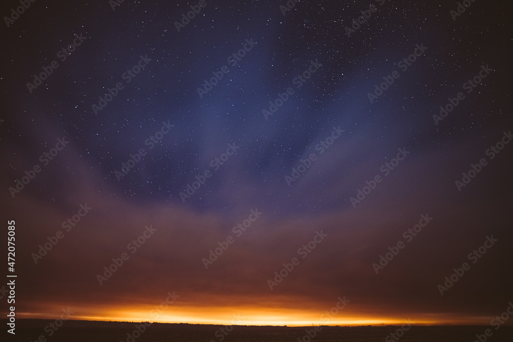 Night Starry Sky With Glowing Stars Above Countryside Landscape. Light Cloudiness Overcast Above Rural Field Meadow In Early Spring. Illumination Lights Of Town On Horizon