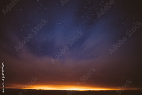 Night Starry Sky With Glowing Stars Above Countryside Landscape. Light Cloudiness Overcast Above Rural Field Meadow In Early Spring. Illumination Lights Of Town On Horizon