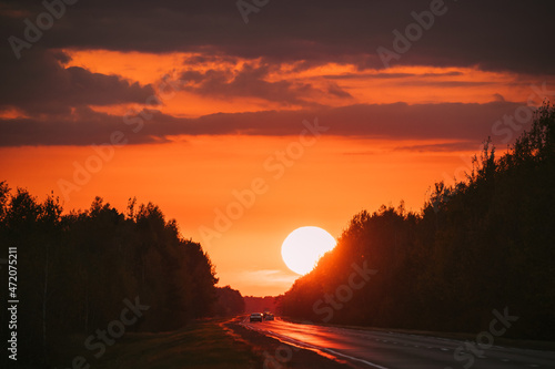 Cars In Motion On Road, Freeway. Asphalt Motorway Highway Against Background Of Big Sunset Sun. Travel Trip Concept. Sunshine Above Road © Grigory Bruev
