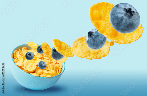 dry cornflakes isolate with blueberries in a blue ceramic plate, part of the cornflakes flies in the air along with the berries. traditional cornflakes breakfast concept