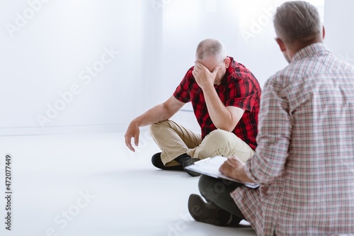 Senior man with mental problems sitting on the floor during counseling session, copy space on empty white wall