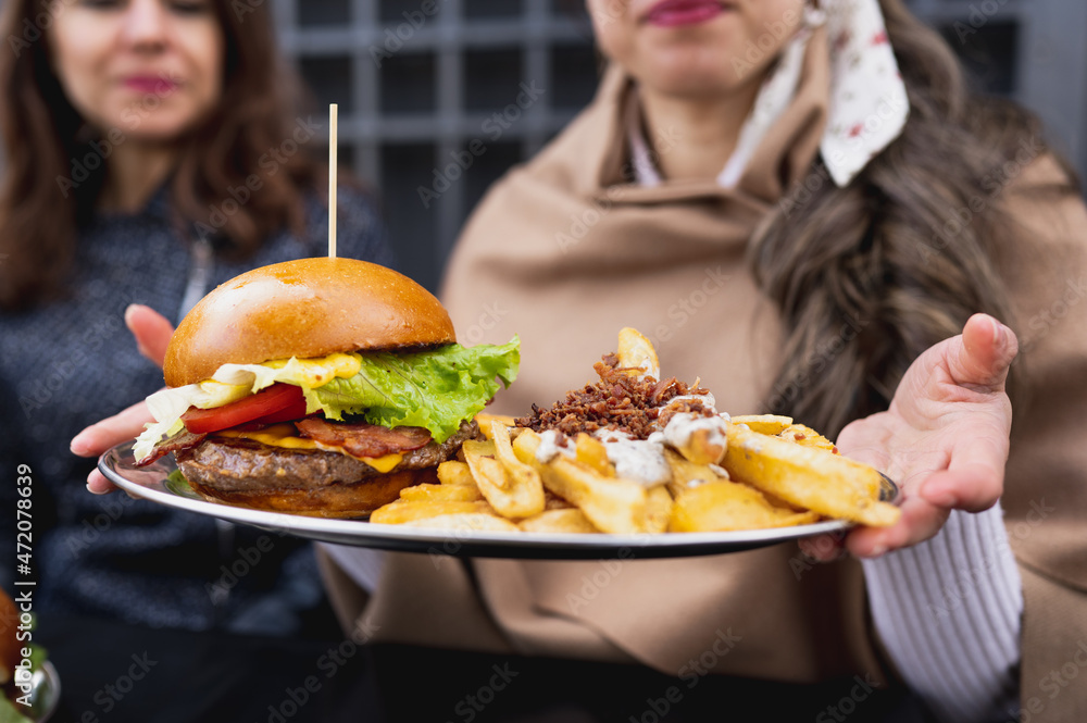 Close-up cropped woman showing plate of gourmet bacon burger with french fries and sauce. Unrecognizable females on background Close-up cropped woman showing plate of gourmet bacon burger with french fries and sauce. Unrecognizable females on background