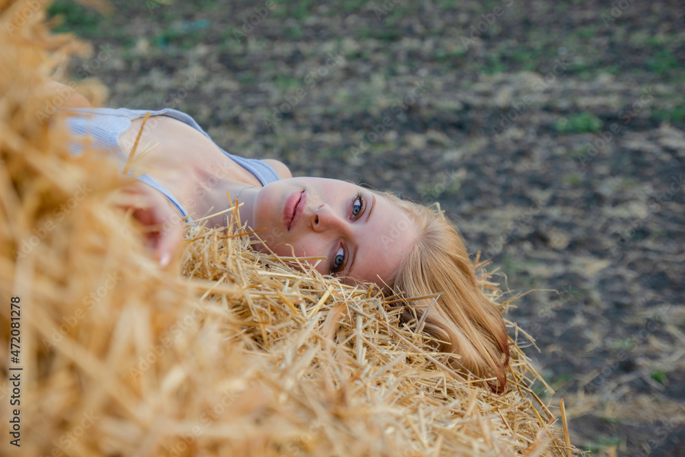 A young beautiful blonde girl with blue eyes and snow-white skin sits on the ground, leaning on a haystack, looks relaxed to the side enjoying the rays of the setting sun in summer. A young beautiful blonde girl with blue eyes and snow-white skin sits on the ground, leaning on a haystack, looks relaxed to the side enjoying the rays of the setting sun in summer.