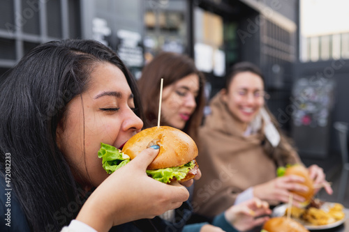 Side view young woman biting a burger. Females sitting at table in restaurant.