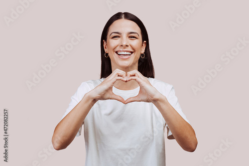 Close up portrait of beautiful caucasian young woman doing love hearth sign with hands at the camera at studio over beige background. Joyful happiness people concept.