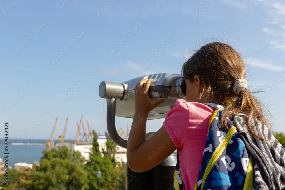 A child girl with a backpack on her back looks through old binoculars to explore the port city on the observation deck in the city park