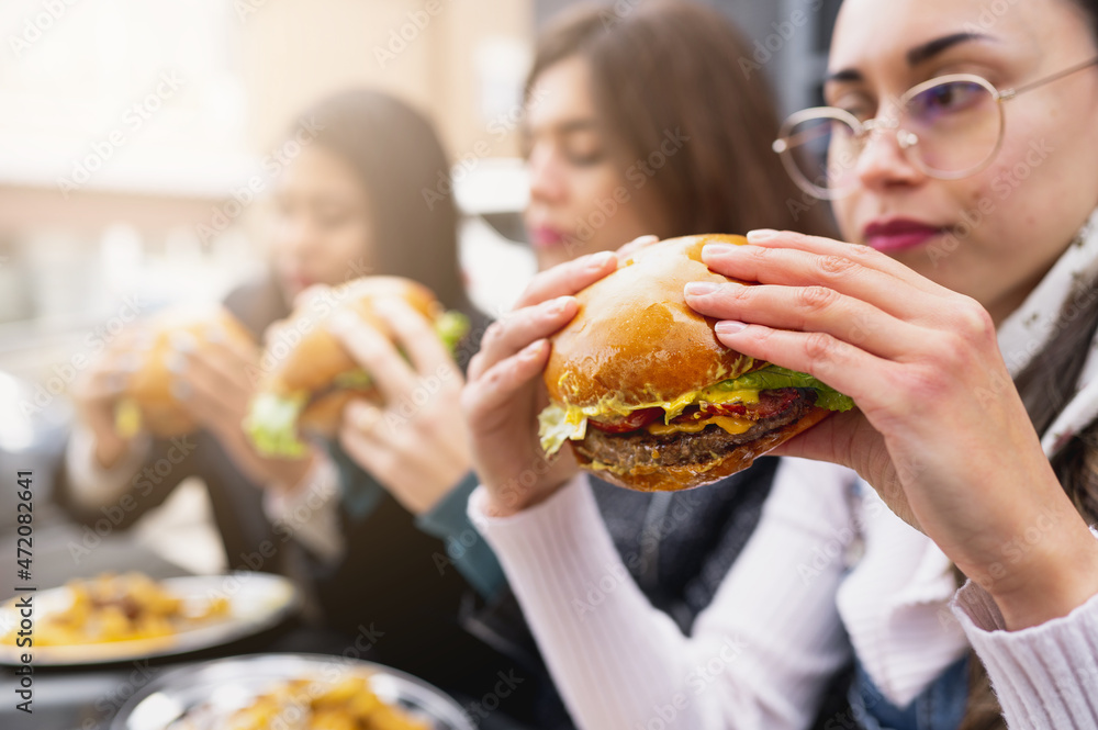 SIde view three young women sitting at table eating burgers. Close-up holding hamburger and bite it. SIde view three young women sitting at table eating burgers. Close-up holding hamburger and bite it.