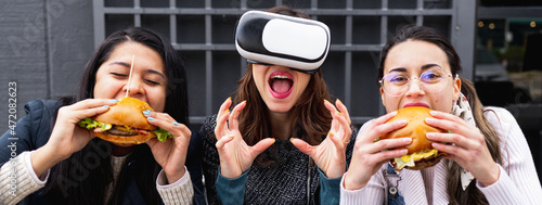 Horizontal banner or header portrait with young women sitting at restaurant eating burgers.  woman wears virtual reality glasses and bite a fictional hamburger. Real life versus virtual concept.