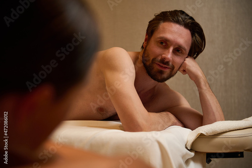 Handsome middle aged European man lying on a massage table, smiles looking at camera, getting ready to receive professional body massage therapy at wellness center. Newlyweds couple at health spa