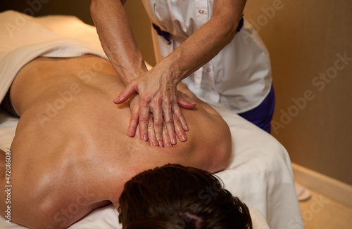 Focus on the hands in massage oil of a physiotherapist massaging the man's back in wellness spa clinic. Unrecognizable man lying on a massage table and enjoying, getting relaxing massage. Body care
