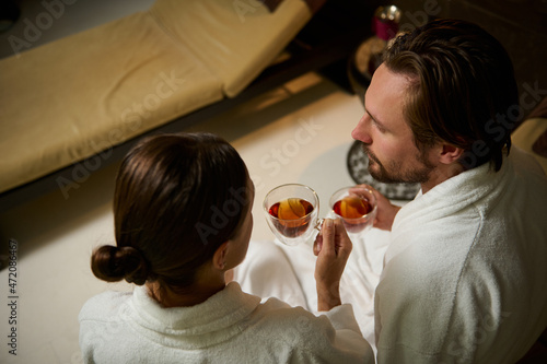 Overhead view of a Caucasian middle aged newlywed, married couple in bathrobes sitting face to face and looking at each other, drinking healthy herbal tea while relaxing in luxury health spa resort