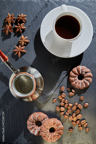 A cup of morning coffee on a white saucer, a coffee maker, cookies, coffee beans, star anise on a black stone stand.