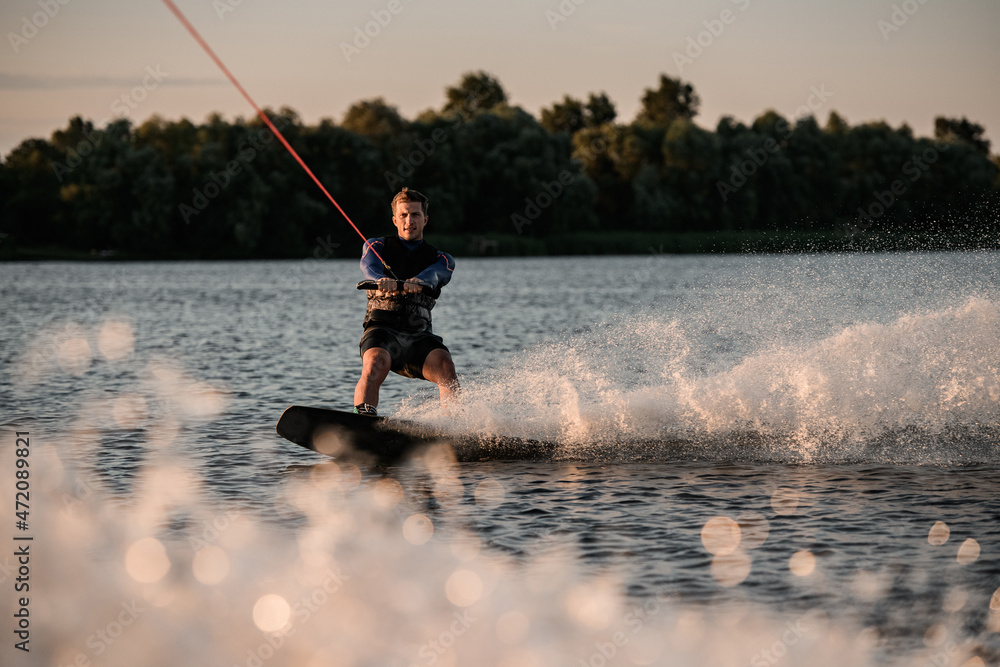 Fototapeta premium Great view of attractive man holding rope and energetically riding wakeboard on splashing river wave.