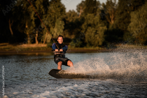 Fototapeta Naklejka Na Ścianę i Meble -  Athletic guy holds rope and speedly riding wakeboard on splashing water surface. Active and extreme sports