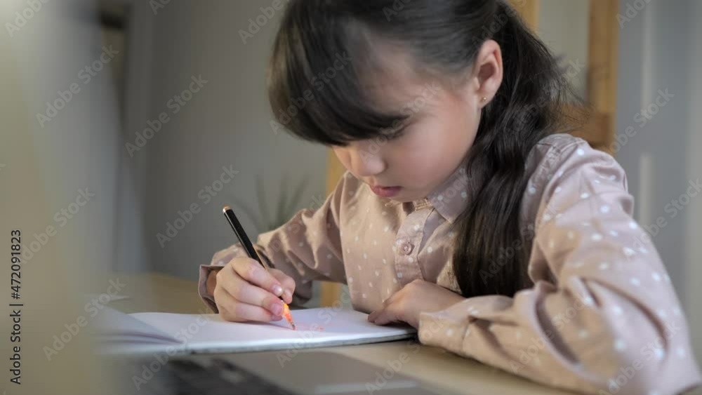 Little girl is writing the essay in notebook at home with laptop on desk. Online learning, homeschooling. Kid is doing homework concept Little girl is writing the essay in notebook at home with laptop on desk. Online learning, homeschooling. Kid is doing homework concept