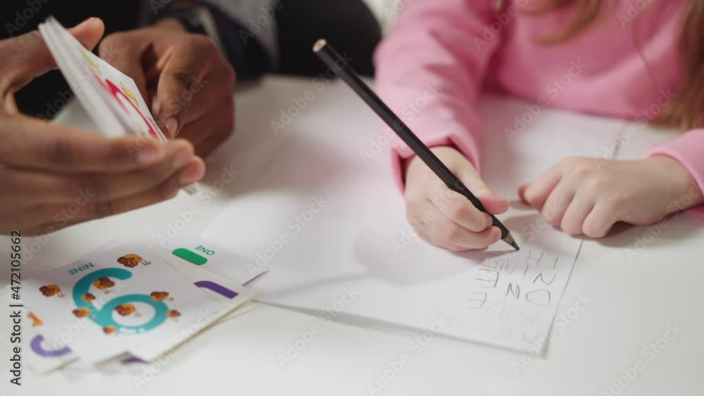 Black English teacher native speaker holds card and little schoolgirl writes digit name on paper at private lesson closeup slow motion Black English teacher native speaker holds card and little schoolgirl writes digit name on paper at private lesson closeup slow motion