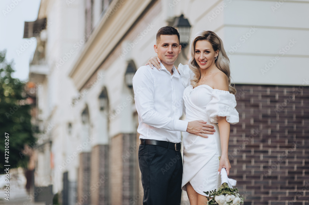 Fototapeta premium A smiling, stylish groom and a beautiful blonde bride in a white dress are hugging on the street in the city against the background of ancient buildings. Wedding photo of newlyweds in love.