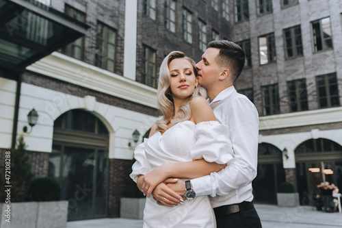 A smiling young groom and a beautiful blonde bride in a white dress are embracing on the street in the city against the background of modern, ancient buildings. Wedding photography of the newlyweds.