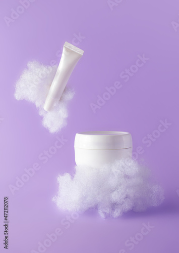 Facial cream and lip balm with fluffy clouds on violet background. Concept care with sensitive skin. Creative still-life photo, vertical. Template white plastic mockups beauty products