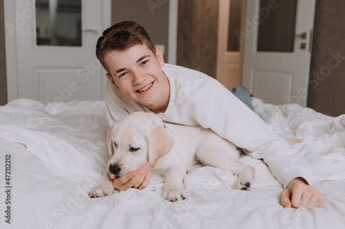 portrait of a teenage boy lying in bed on white bedding in an embrace with a light-colored dog. boy is fooling around with a pet in the room. space for text. High quality photo