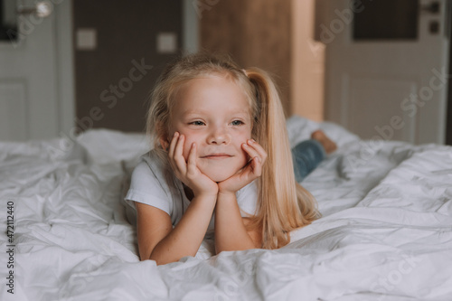 portrait of little smiling blonde girl in a white T-shirt and jeans lying on her stomach in bed with white bedding. children's recreation. space for text. High quality photo