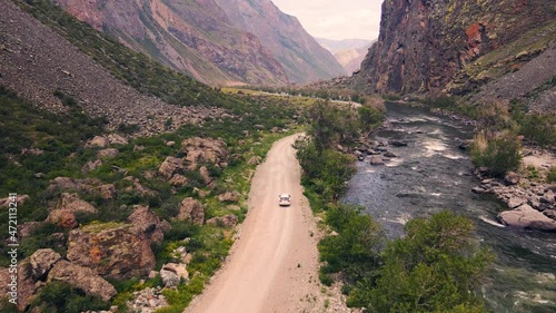 A white SUV jeep car is driving along an empty country road in the huge Katu-Yaryk canyon Chulyshman Valley with a large mountain bubbling river. Altai, Siberia, Russia. People travel along a dirt photo