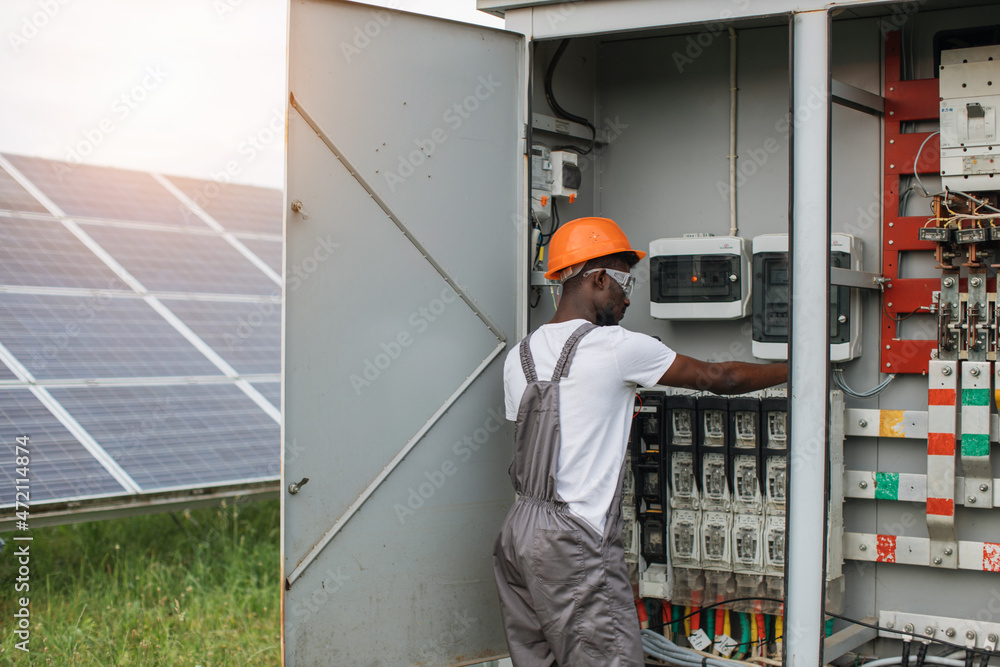 African american man in grey overalls and orange helmet repairing cables in switchgear at solar station. Concept of people, maintenance and alternative energy. African american man in grey overalls and orange helmet repairing cables in switchgear at solar station. Concept of people, maintenance and alternative energy.