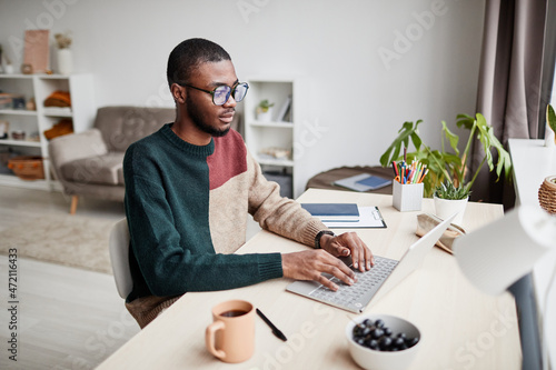 Portrait of young African-American man wearing glasses while working from home and using laptop, copy space photo