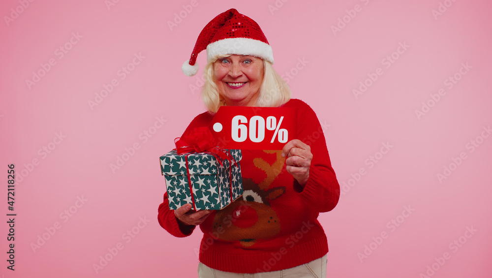 Grandmother woman in Christmas sweater showing gift box and 60 Percent discount inscriptions banner text. Black Friday. Good holiday low price for shopping. Blue studio wall background. Happy New Year