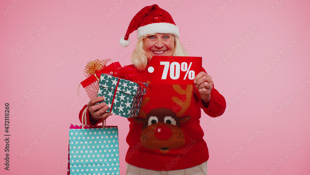 Obraz premium Grandmother old woman in Christmas sweater showing gift box and 70 Percent discount inscriptions banner text. Black Friday. Good holiday low prices for shopping. Blue studio background. Happy New Year
