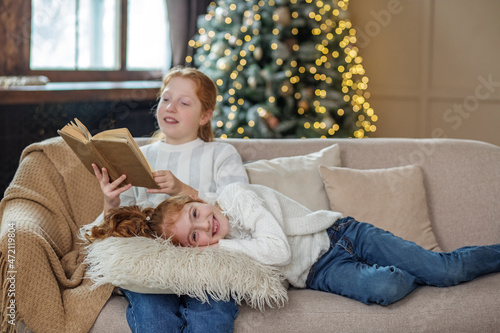 Two sisters are sitting on the couch and reading Christmas stories. Delicious hot cocoa.
