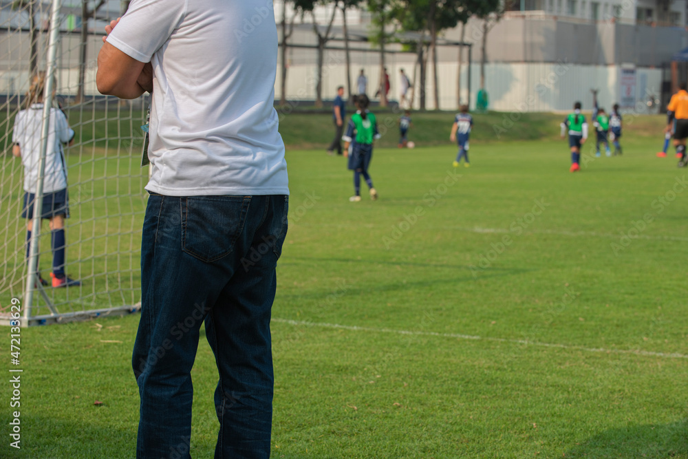 Father standing and watching his daughter playing football in a school tournament on a sideline with a sunny day. Sport, outdoor active, lifestyle, happy family and soccer mom and soccer dad concepts.