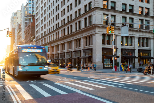 Busy street scene with people, bus and taxi at the intersection of 23rd and 5th Avenue in New York City with sunlight background