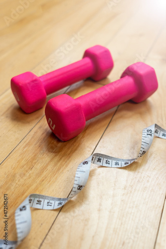 A white measuring tape laying next to two small pink dumbbells on the wooden floor of a home gym
