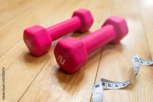 Two small pink dumbbells laying on the floor of a home gym next to a white measuring tape