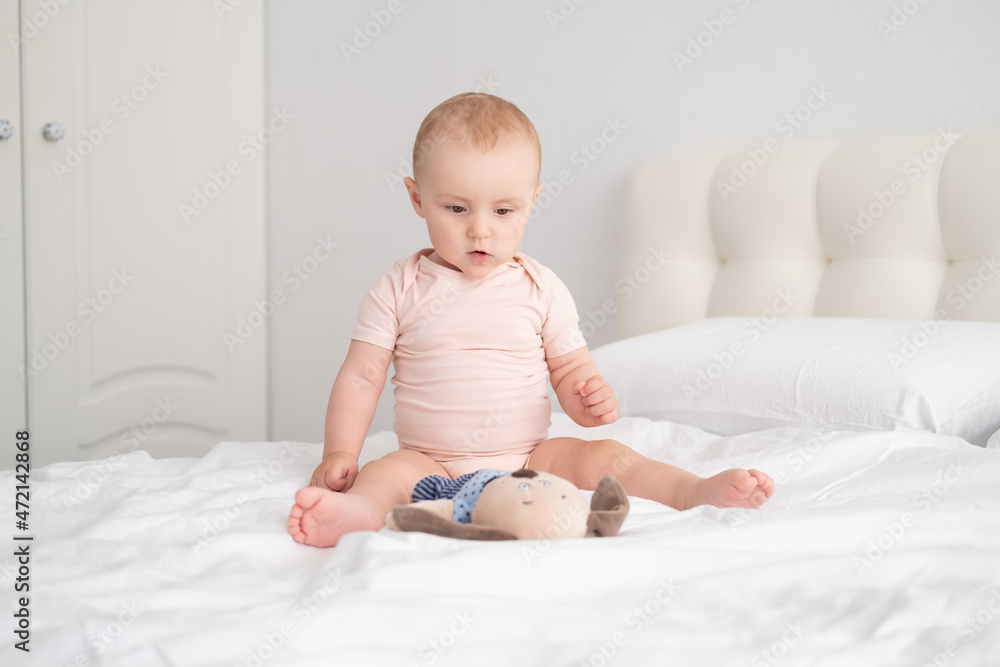 baby girl in light pink bodysuit playing with soft toy on white bedding on bed