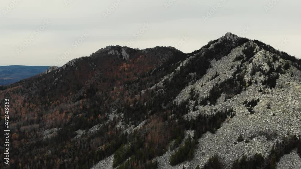 Aerial view of the top of the mountain from pile of stones in the autumn forest. Flying over green and gray trees. Other mountain peaks can be seen in the distance. Late autumn. Drone shooting Aerial view of the top of the mountain from pile of stones in the autumn forest. Flying over green and gray trees. Other mountain peaks can be seen in the distance. Late autumn. Drone shooting